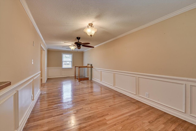 empty room featuring a textured ceiling, a decorative wall, a ceiling fan, light wood-style floors, and ornamental molding