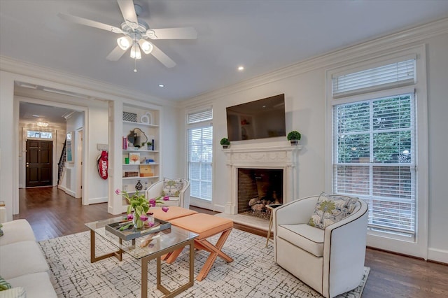 living area featuring built in features, a ceiling fan, wood finished floors, a fireplace with raised hearth, and ornamental molding