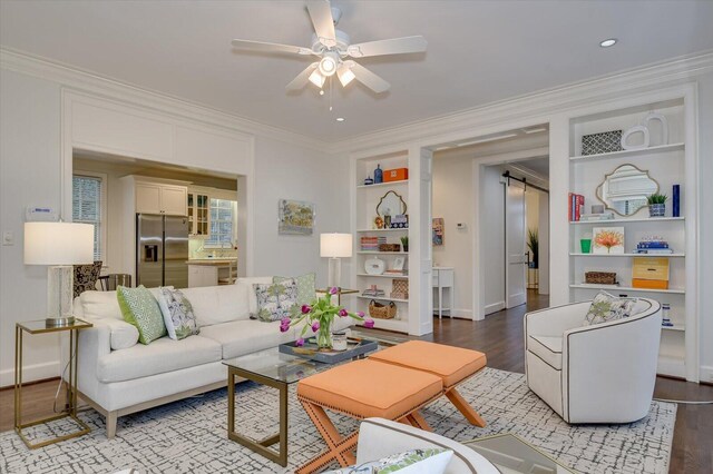 living area featuring built in shelves, a ceiling fan, wood finished floors, ornamental molding, and a barn door