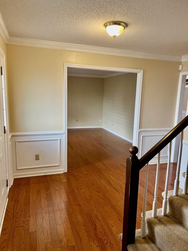unfurnished room featuring wood finished floors, crown molding, a textured ceiling, a wainscoted wall, and stairs