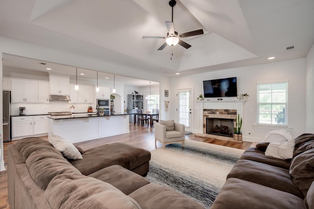 living room featuring ceiling fan, a fireplace, light wood finished floors, visible vents, and a tray ceiling