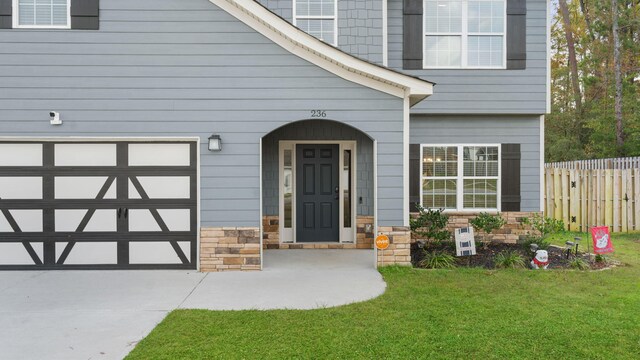 doorway to property with fence, concrete driveway, a garage, stone siding, and a lawn