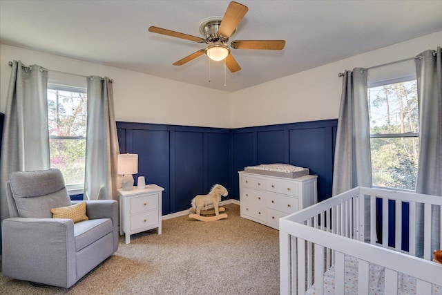 carpeted bedroom featuring a wainscoted wall, a crib, a decorative wall, and ceiling fan