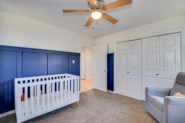 carpeted bedroom featuring a crib, ceiling fan, wainscoting, a closet, and a decorative wall