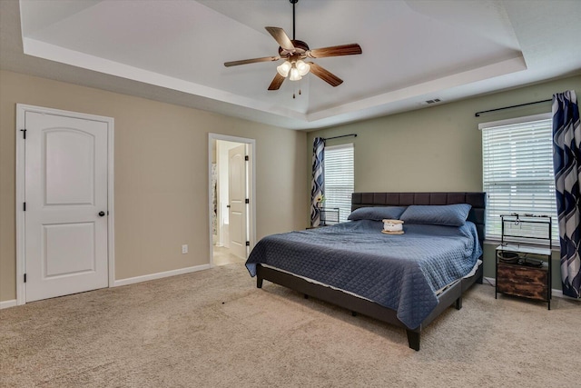 bedroom featuring light carpet, ensuite bathroom, ceiling fan, a tray ceiling, and multiple windows