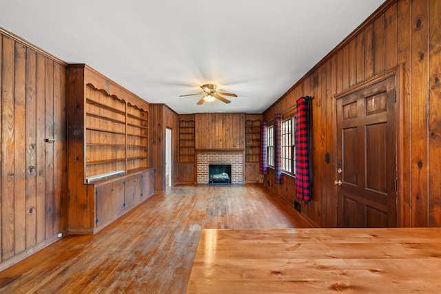 unfurnished living room with built in shelves, a brick fireplace, wooden walls, ceiling fan, and hardwood / wood-style floors