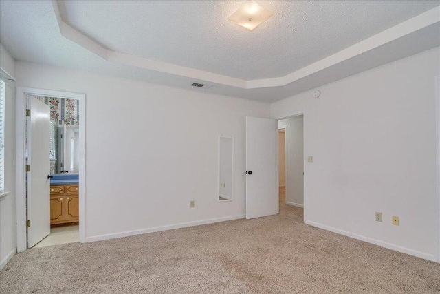 unfurnished bedroom featuring light carpet, a textured ceiling, a tray ceiling, and ensuite bathroom