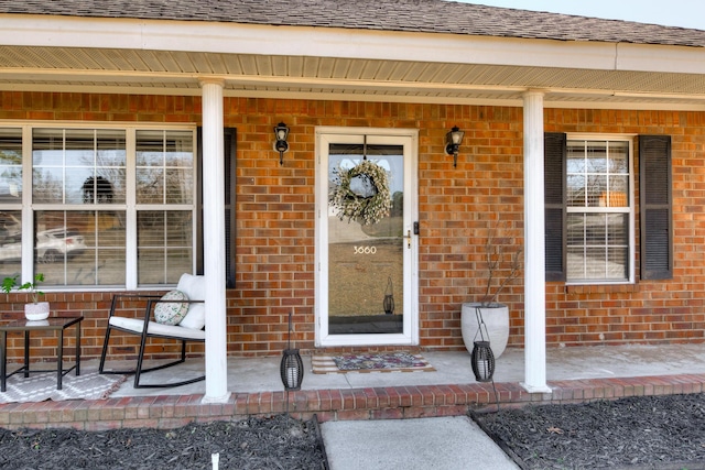 view of exterior entry with covered porch, a shingled roof, and brick siding