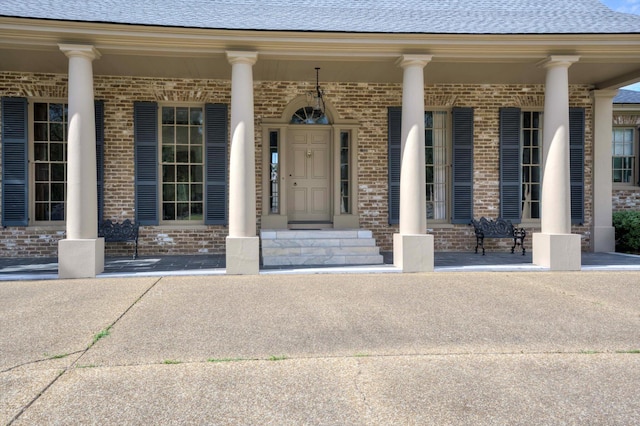 view of exterior entry with covered porch, brick siding, and roof with shingles