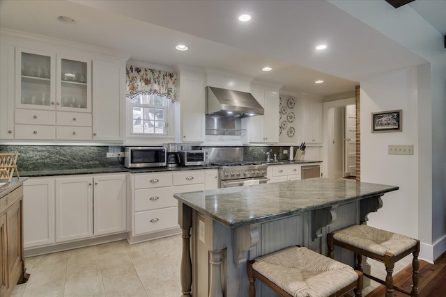 kitchen featuring decorative backsplash, appliances with stainless steel finishes, a kitchen breakfast bar, wall chimney exhaust hood, and white cabinets