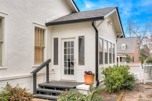 property entrance with stucco siding, roof with shingles, and fence