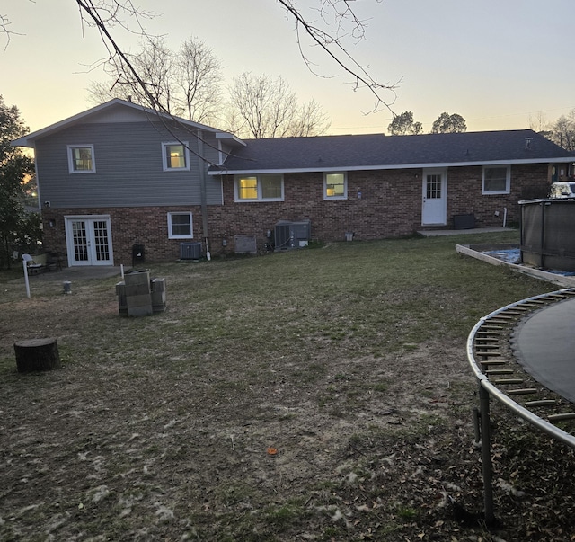 back of property at dusk with a yard, french doors, brick siding, and central air condition unit