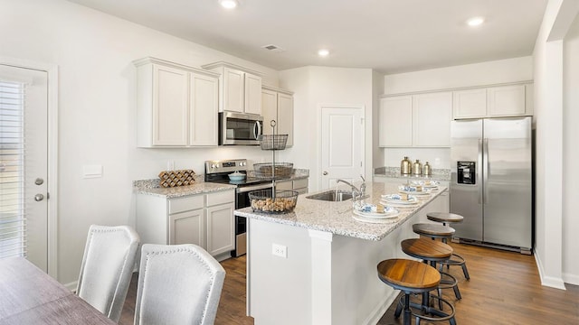 kitchen with appliances with stainless steel finishes, a kitchen breakfast bar, light stone counters, a center island with sink, and dark hardwood / wood-style flooring