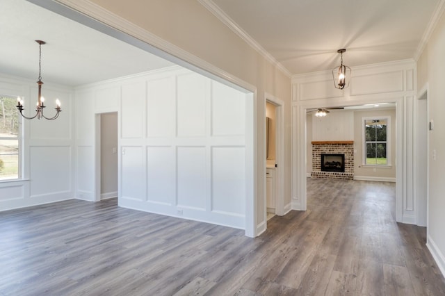 unfurnished dining area with wood finished floors, a brick fireplace, crown molding, a decorative wall, and ceiling fan with notable chandelier