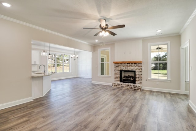 unfurnished living room featuring a brick fireplace, wood finished floors, crown molding, and ceiling fan with notable chandelier