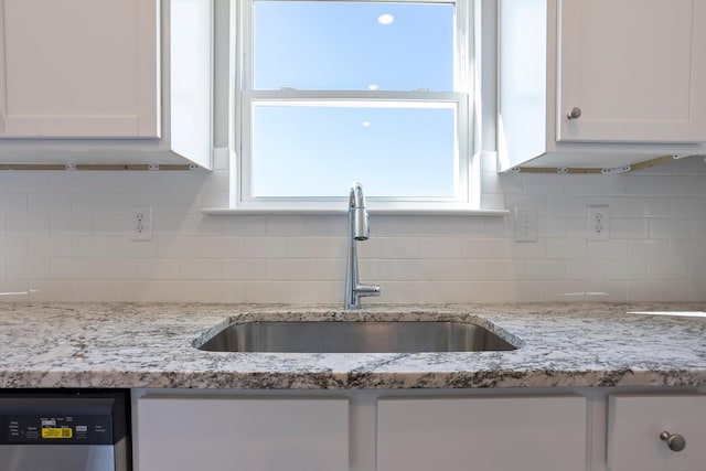 kitchen with light stone counters, backsplash, stainless steel dishwasher, white cabinetry, and a sink