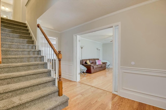 staircase featuring crown molding, ceiling fan, a wainscoted wall, wood finished floors, and a textured ceiling