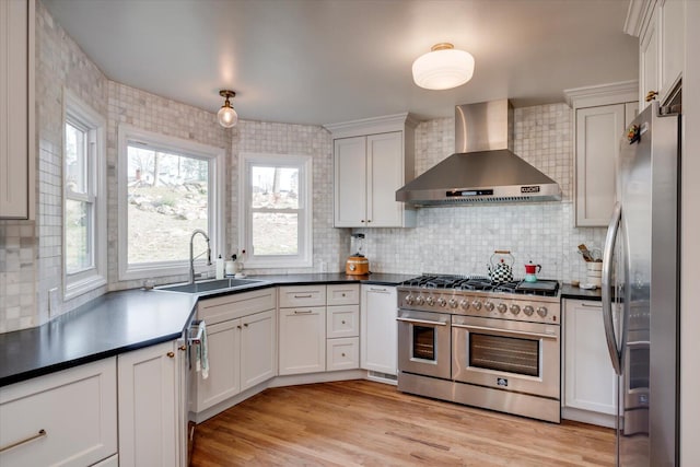 kitchen featuring stainless steel appliances, dark countertops, light wood-style floors, a sink, and wall chimney range hood