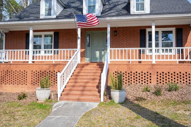 entrance to property with a shingled roof, covered porch, and brick siding