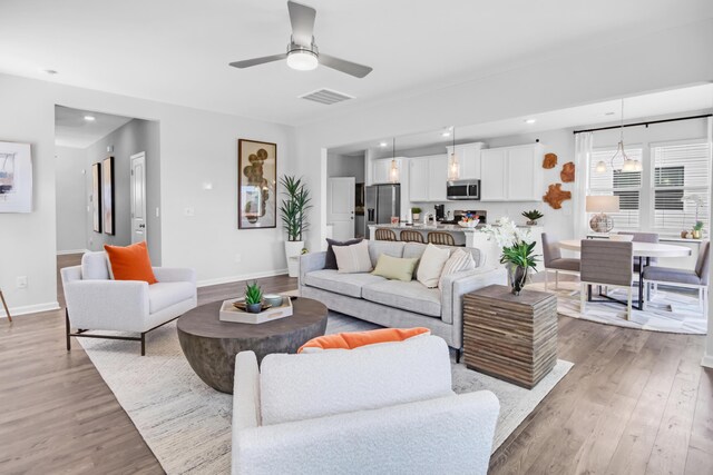 living room featuring light wood-type flooring, visible vents, baseboards, and ceiling fan with notable chandelier