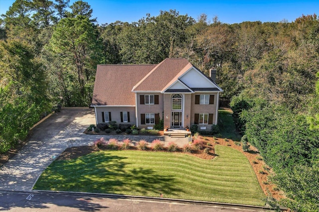 colonial inspired home featuring a forest view, a chimney, a front lawn, and concrete driveway