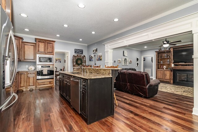 kitchen with appliances with stainless steel finishes, light stone counters, ceiling fan, dark hardwood / wood-style floors, and an island with sink