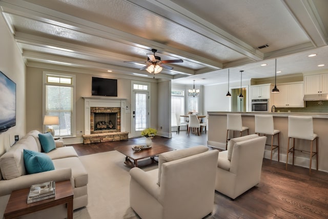 living room featuring ceiling fan with notable chandelier, crown molding, beamed ceiling, dark hardwood / wood-style floors, and a stone fireplace