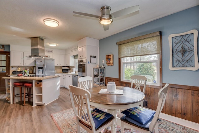 dining room featuring wooden walls, wainscoting, wood finished floors, a textured ceiling, and a ceiling fan