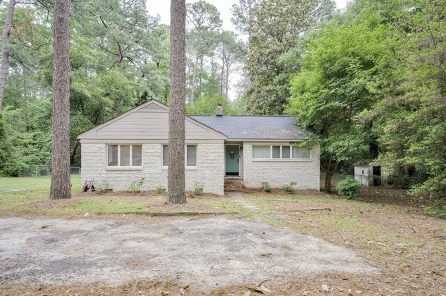 ranch-style house with brick siding, a chimney, roof with shingles, view of wooded area, and fence