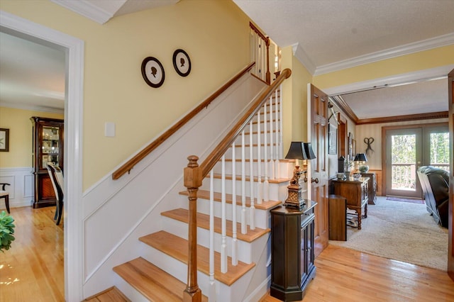 stairway featuring wood finished floors, french doors, a decorative wall, crown molding, and wainscoting