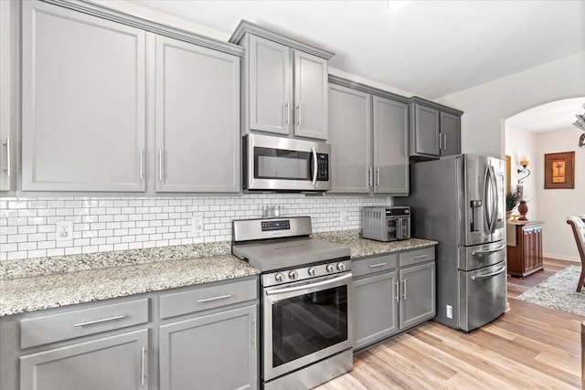 kitchen featuring gray cabinetry, light stone countertops, light hardwood / wood-style floors, decorative backsplash, and appliances with stainless steel finishes