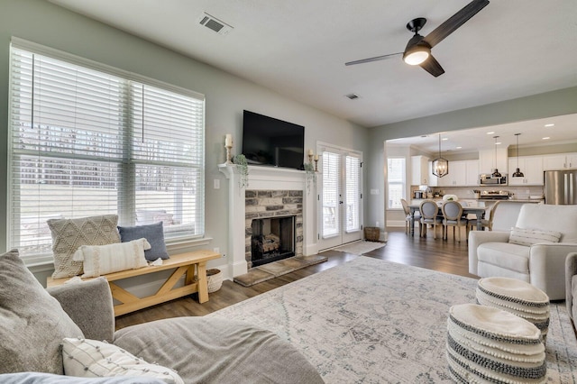 living area featuring a stone fireplace, wood finished floors, visible vents, and ceiling fan