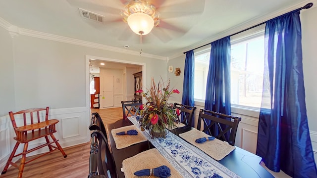 dining room with a ceiling fan, wood finished floors, visible vents, ornamental molding, and wainscoting