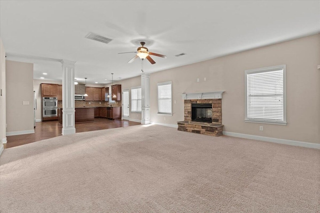 unfurnished living room featuring ceiling fan, dark carpet, a stone fireplace, and decorative columns