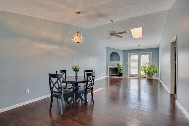 dining space with a textured ceiling, dark wood-style flooring, baseboards, lofted ceiling with skylight, and a glass covered fireplace