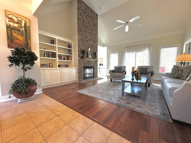 tiled living room featuring a stone fireplace, ceiling fan, built in shelves, and high vaulted ceiling