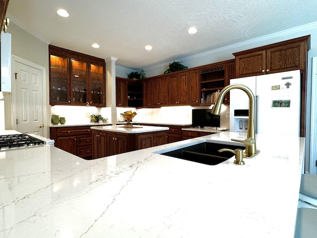 kitchen featuring sink, light stone counters, white refrigerator with ice dispenser, a textured ceiling, and ornamental molding