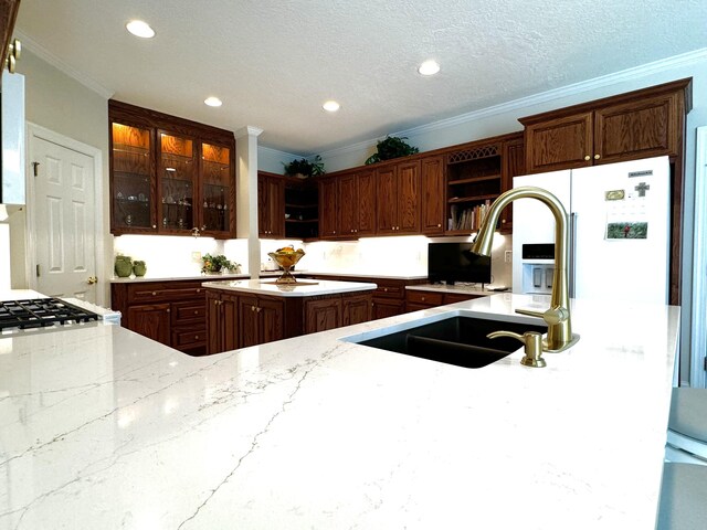 kitchen featuring sink, light stone counters, white refrigerator with ice dispenser, a textured ceiling, and ornamental molding