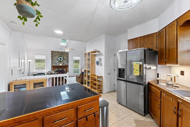 kitchen featuring ceiling fan, a stone fireplace, stainless steel refrigerator with ice dispenser, backsplash, and light tile patterned floors