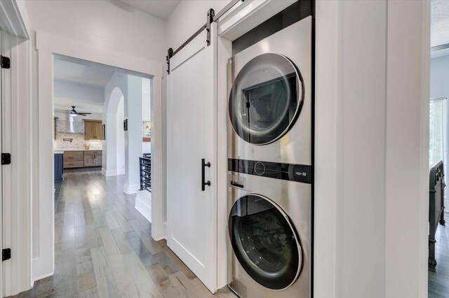 laundry room with ceiling fan, a barn door, stacked washer / dryer, and light hardwood / wood-style flooring