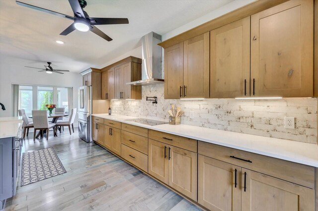 kitchen with wall chimney range hood, light hardwood / wood-style flooring, ceiling fan, black electric cooktop, and tasteful backsplash