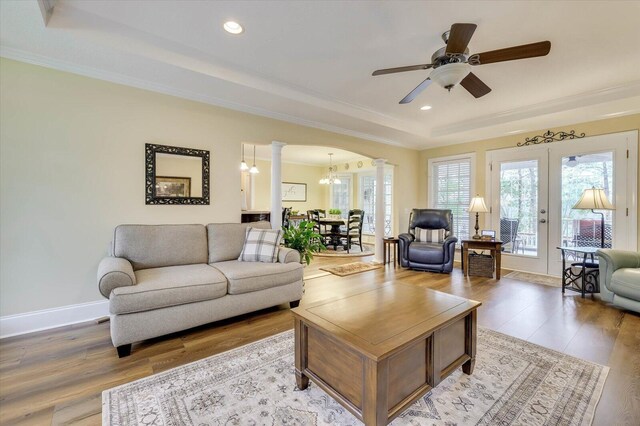 living room with a raised ceiling, ornate columns, ceiling fan with notable chandelier, and light wood-type flooring