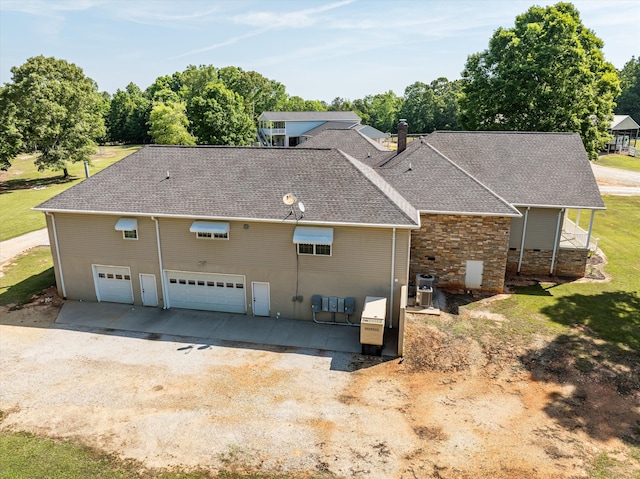 back of house featuring an attached garage, driveway, roof with shingles, central AC unit, and view of wooded area