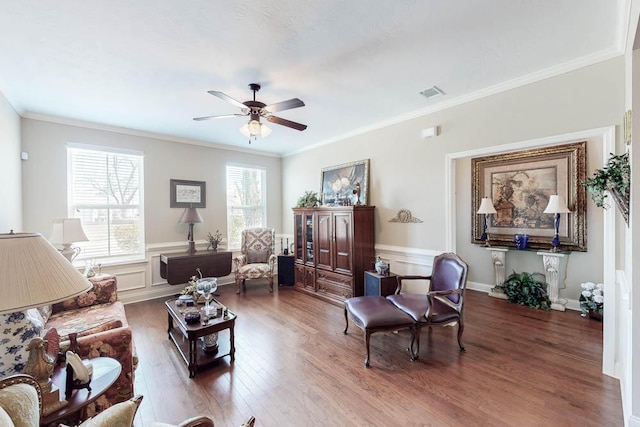 living room with crown molding, visible vents, dark wood-type flooring, wainscoting, and ceiling fan