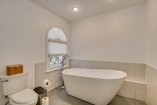 bathroom with toilet, a soaking tub, tile walls, and a wainscoted wall