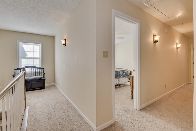 corridor with attic access, baseboards, a textured ceiling, and light colored carpet