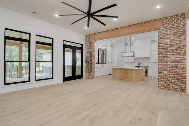 unfurnished living room with ceiling fan with notable chandelier, french doors, light wood-type flooring, and brick wall