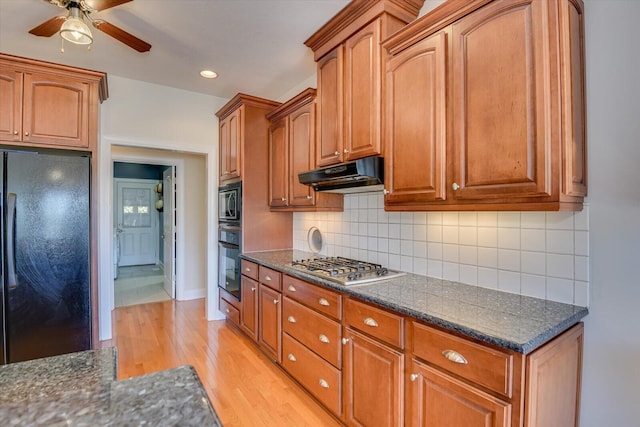 kitchen with tasteful backsplash, light wood-style flooring, dark stone countertops, under cabinet range hood, and black appliances