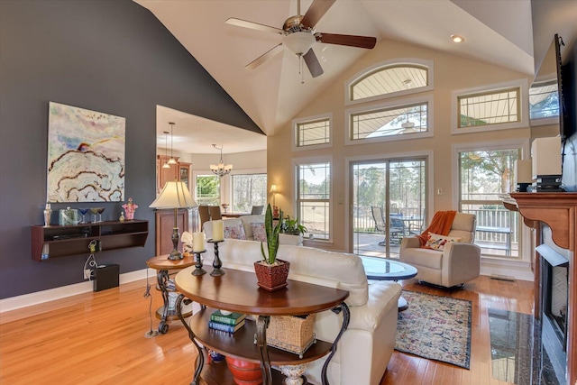 living area with high vaulted ceiling, ceiling fan with notable chandelier, a fireplace with flush hearth, baseboards, and light wood-style floors