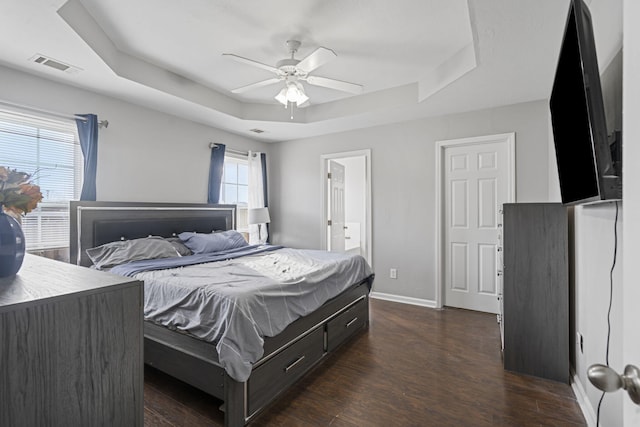 bedroom with dark wood-type flooring, ceiling fan, ensuite bathroom, and a tray ceiling